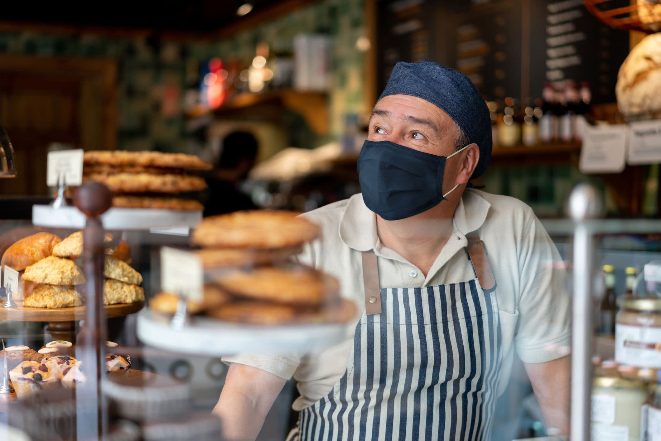 Mitarbeiter in Bäckerei mit Maske – lohnrelevante Arbeitszeit im Verkauf Ein Bäckereimitarbeiter mit gestreifter Schürze und Maske steht hinter der Verkaufstheke – lohnbuchhalterisch relevant im Hinblick auf Arbeitszeiten und Mindestlohn