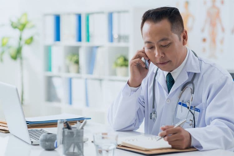 Arzt telefoniert am Schreibtisch und macht sich Notizen Arzt mit Stethoskop am Schreibtisch telefoniert und notiert Informationen – Symbolbild für Praxisorganisation und Lohnbuchhaltung im Gesundheitswesen