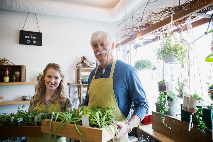 Mitarbeiter in einem Familienbetrieb beim Verkauf von Pflanzen – Lohnbuchhaltung im Kleinunternehmen Zwei Personen mit grünen Schürzen, vermutlich Vater und Tochter, halten Pflanzen in einem hellen Blumenladen oder Gartencenter