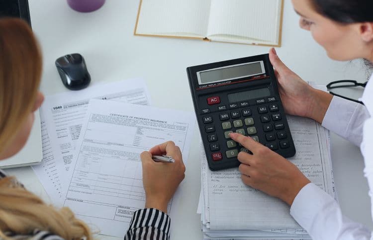 Zwei Personen bei der Berechnung von Zahlen mit einem großen Taschenrechner – Lohnbuchhaltung Zwei Personen sitzen an einem Schreibtisch mit einem großen Taschenrechner und Dokumenten, eine Person tippt, die andere schreibt – Symbolbild für Lohnbuchhaltung und Gehaltsabrechnung