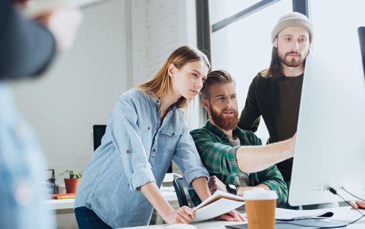 Drei junge Menschen arbeiten gemeinsam an einem großen Monitor im Büro Zwei Männer und eine Frau blicken konzentriert auf einen großen Computerbildschirm. Sie stehen und sitzen gemeinsam an einem modernen Arbeitsplatz mit Notizblock und Kaffeebecher.