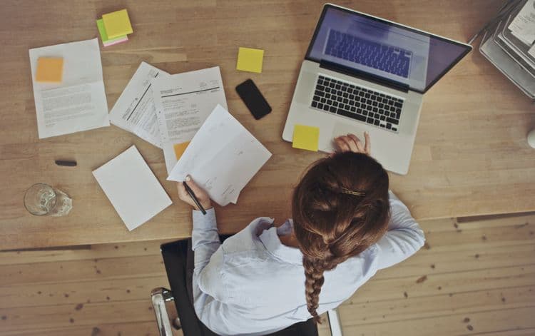 Lohnbuchhaltung im Homeoffice: Mitarbeiterin bei der Bearbeitung von Gehaltsunterlagen Frau mit geflochtenem Zopf arbeitet im Homeoffice an einem Holztisch mit Laptop, Unterlagen und Notizzetteln – symbolisiert Tätigkeiten in der Lohnbuchhaltung