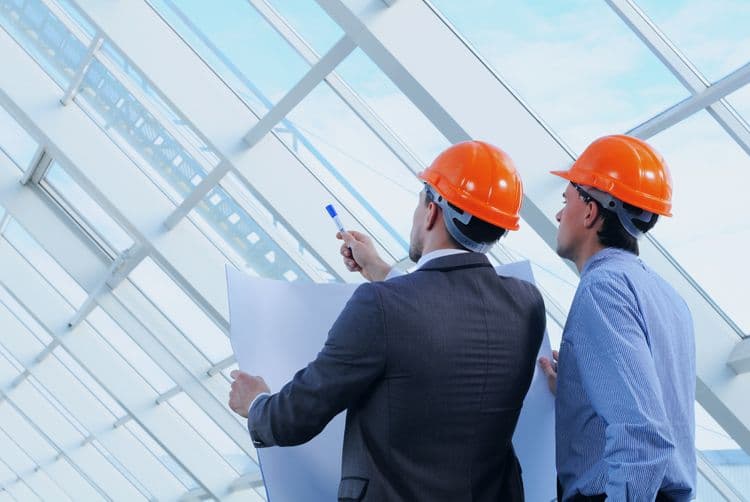 Bauleiter im Gespräch zur Projektplanung – relevante Informationen für die Lohnbuchhaltung im Baugewerbe Zwei Männer mit orangefarbenen Schutzhelmen betrachten gemeinsam einen Bauplan in einer modernen, lichtdurchfluteten Industriehalle
