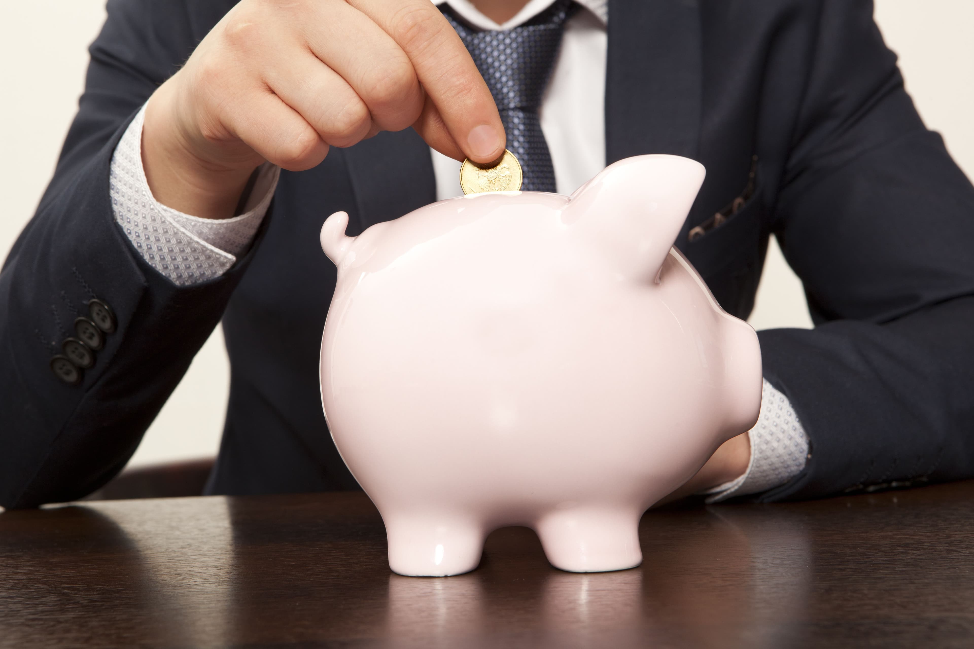 Geschäftsmann wirft eine Münze in ein rosa Sparschwein Ein Mann im Anzug steckt eine Münze in ein rosa Sparschwein – Symbol für Geld sparen, Finanzplanung und Vermögensaufbau