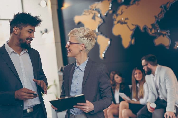 Fachgespräch zur internationalen Lohnbuchhaltung vor Weltkarte im Büro Zwei Personen im Business-Outfit unterhalten sich vor einer Weltkarte im Büro – im Hintergrund weitere Kollegen – symbolisch für internationale Aspekte der Lohnbuchhaltung
