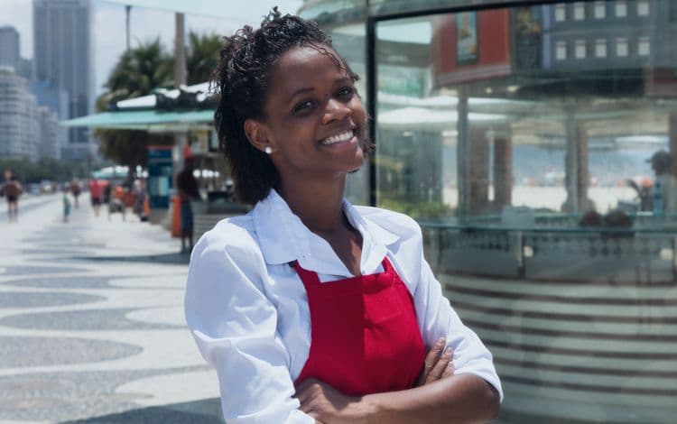 Zufriedene Servicekraft: Lohnbuchhaltung im Gastgewerbe Lächelnde Mitarbeiterin in roter Schürze steht vor einem Restaurant und blickt in die Kamera