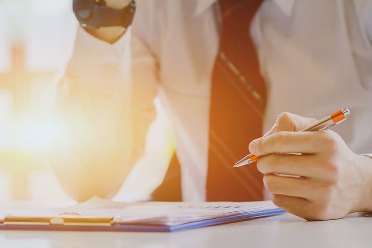 Lohnbuchhaltung: Geschäftsmann prüft Unterlagen am Schreibtisch Nahaufnahme einer männlichen Hand mit Kugelschreiber über einem Klemmbrett mit Dokumenten – symbolisiert die manuelle Prüfung und Dokumentation in der Lohnbuchhaltung